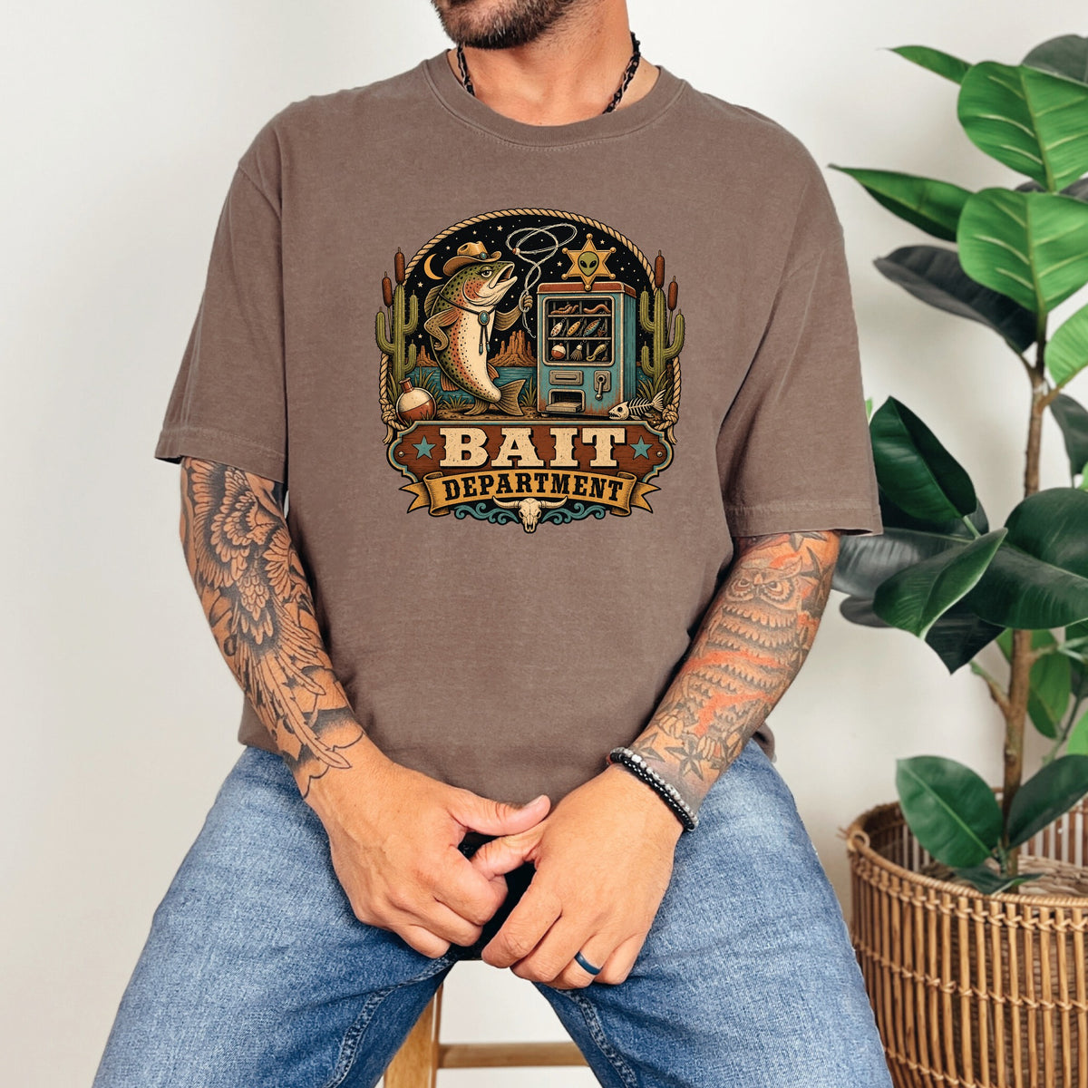 A tattooed man wears a brown t-shirt featuring a vintage Bait Department graphic.