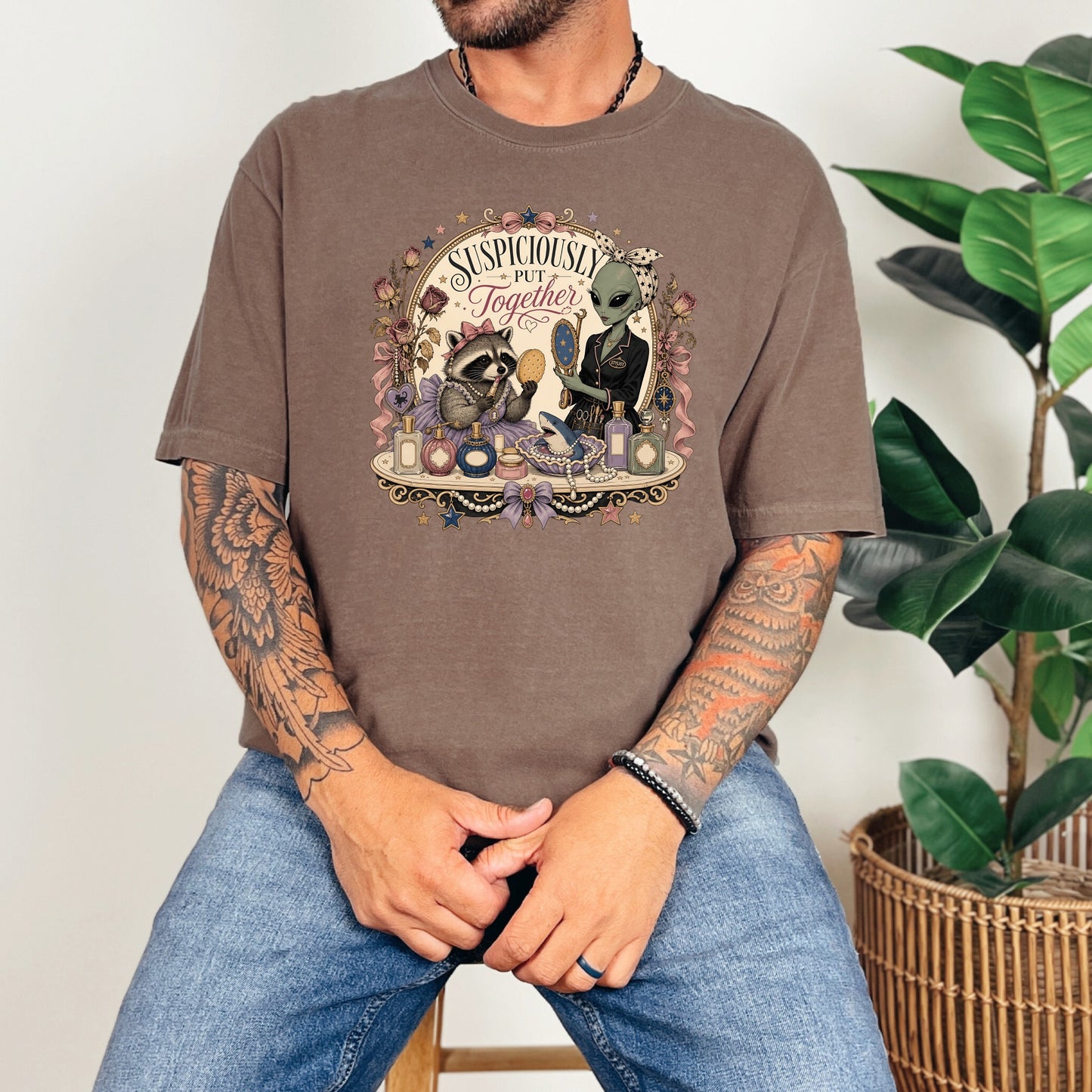 A tattooed man wearing a brown t-shirt featuring a raccoon and alien graphic with the text Suspiciously Put Together.