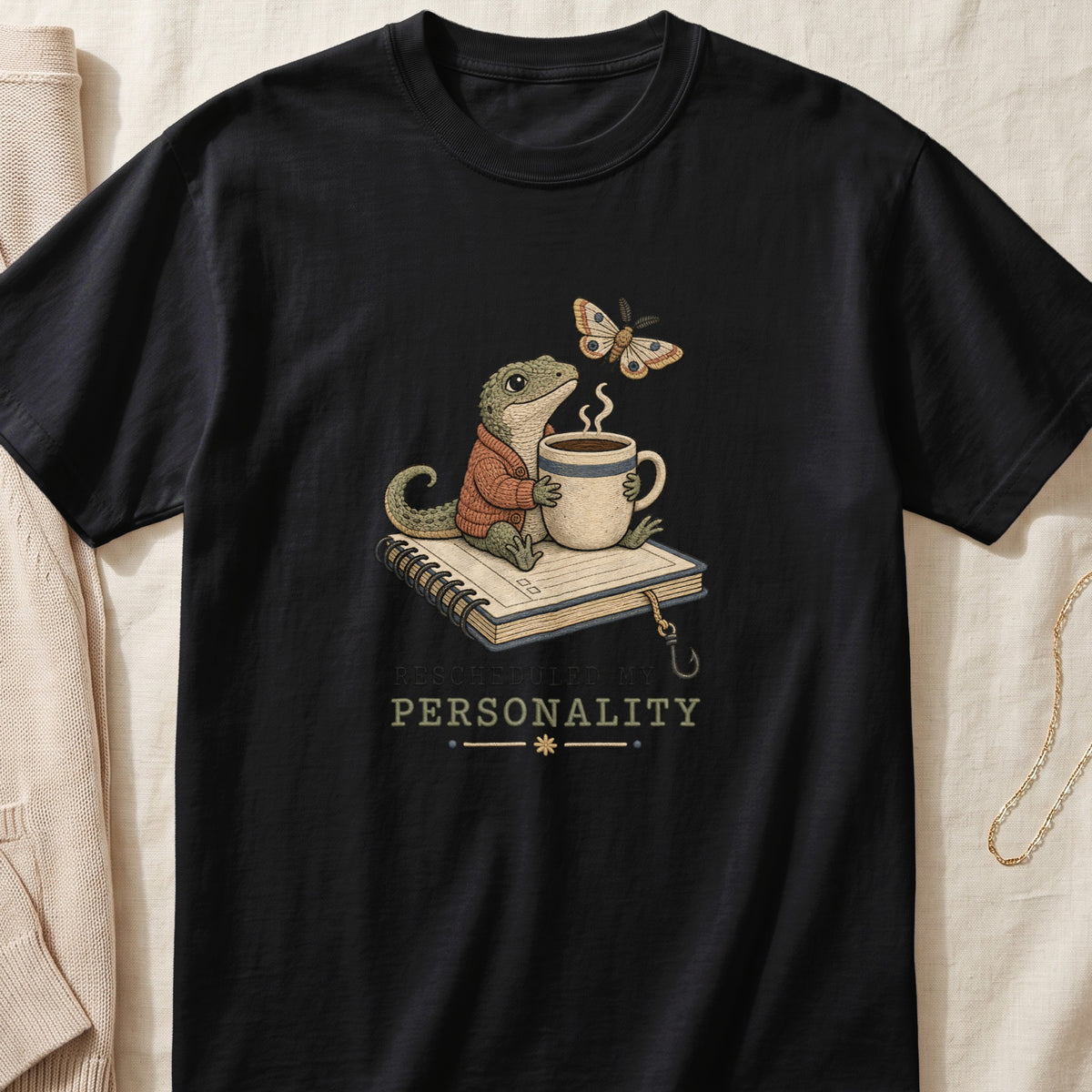 A black t-shirt features a graphic of a gecko wearing a sweater, holding a coffee cup atop a book, with a moth and the text "RESCHEDULED MY PERSONALITY."