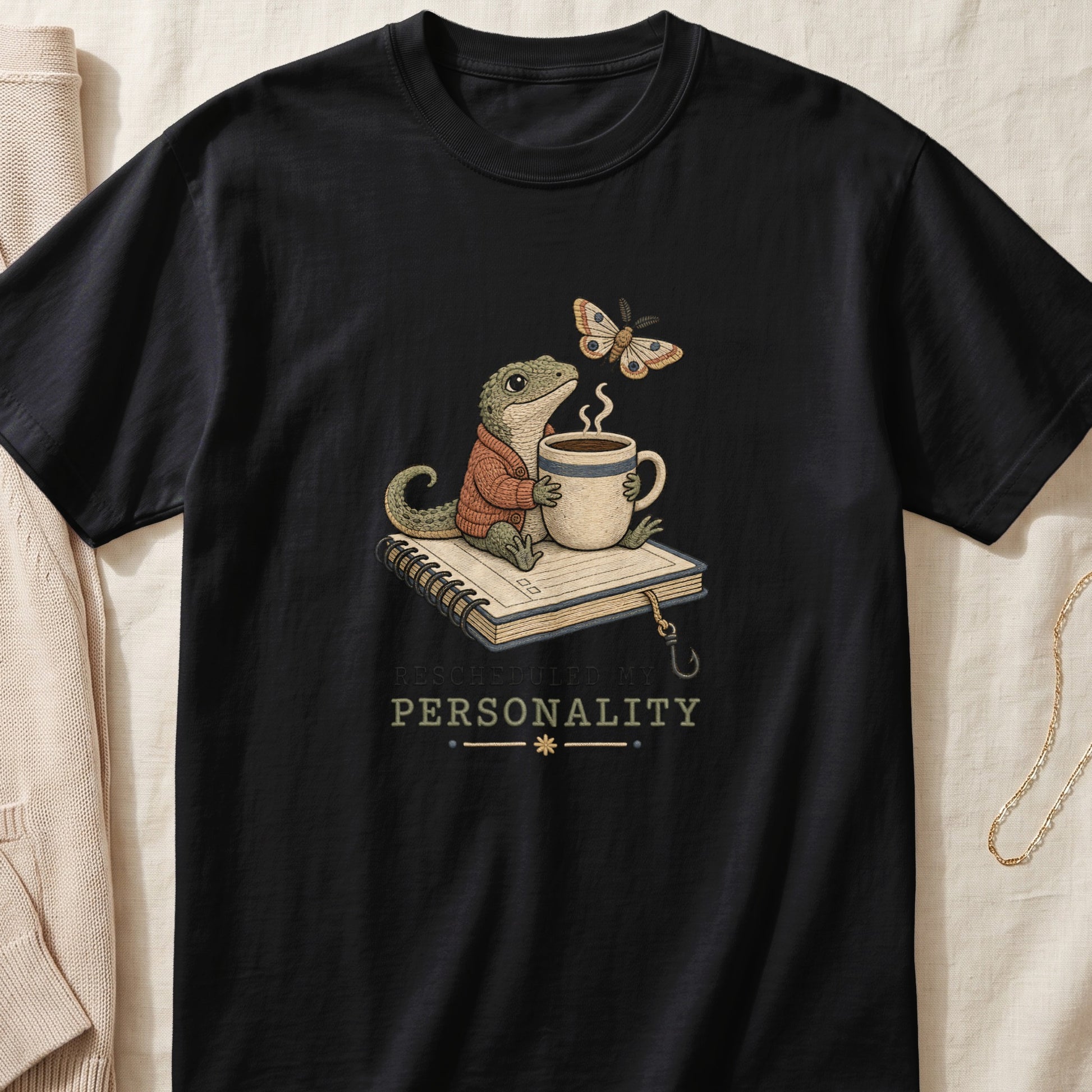 A black t-shirt features a graphic of a gecko wearing a sweater, holding a coffee cup atop a book, with a moth and the text "RESCHEDULED MY PERSONALITY."