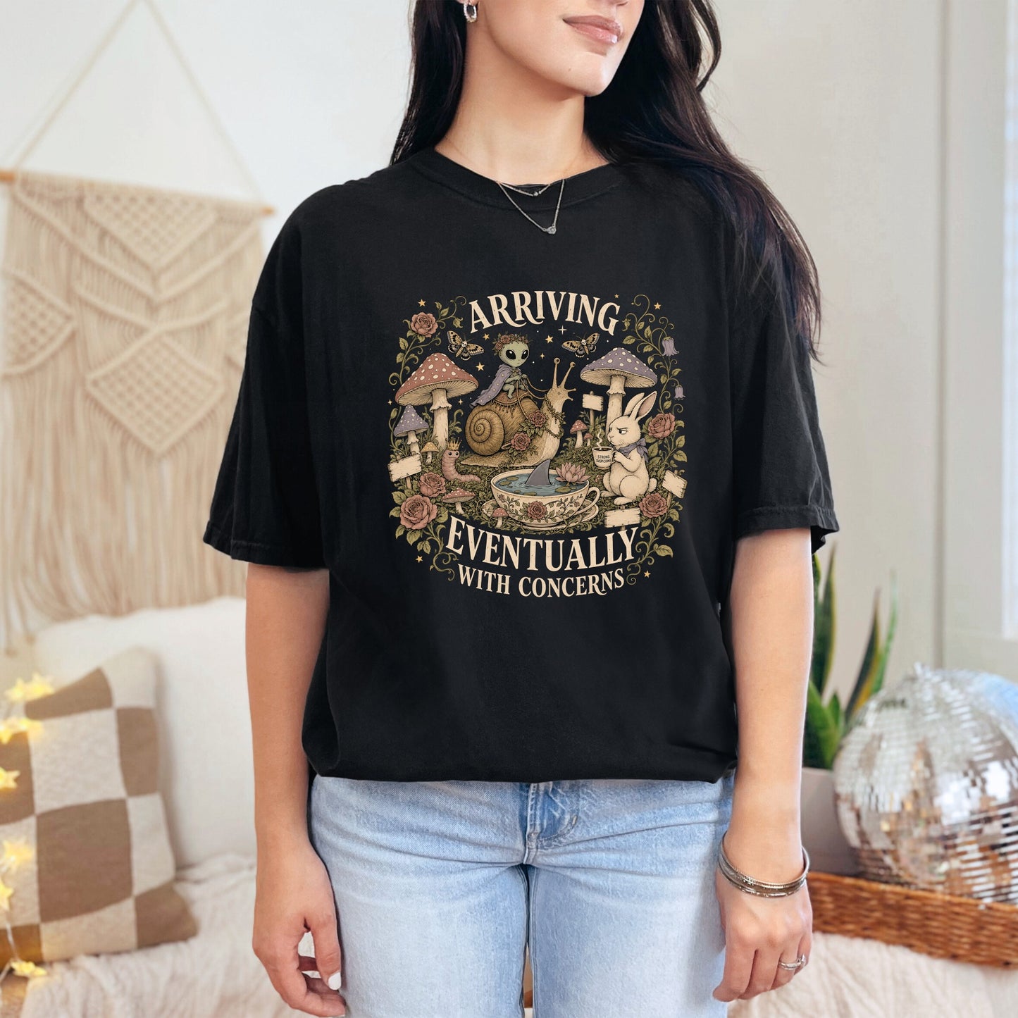 Black t-shirt graphic with alien, snail, rabbit, and mushrooms, reading "Arriving Eventually With Concerns."