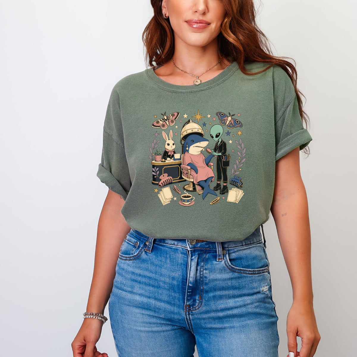 A woman wears an olive green t-shirt featuring a vintage-style graphic of a shark getting its hair styled by an alien, with a rabbit watching.