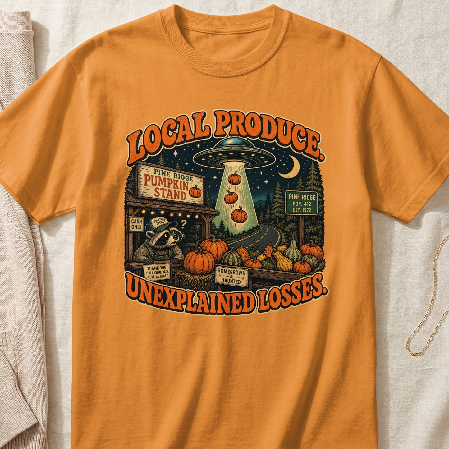 Orange t-shirt graphic with UFO and raccoon at a pumpkin stand, text "Local Produce Unexplained Losses".