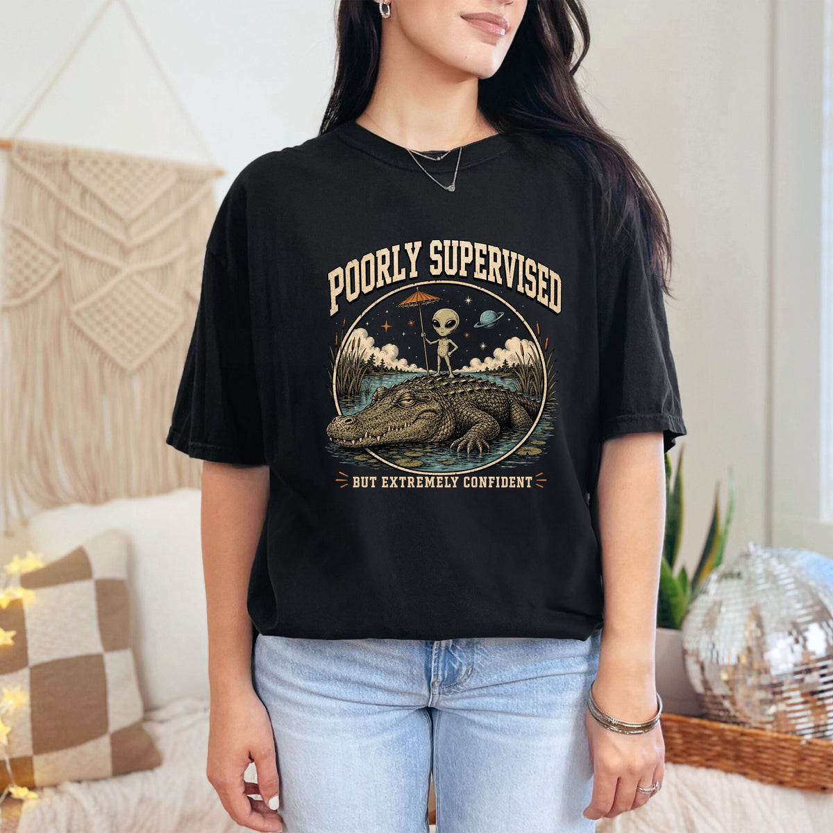 Black t-shirt graphic with alligator, alien holding umbrella, text: "POORLY SUPERVISED BUT EXTREMELY CONFIDENT".