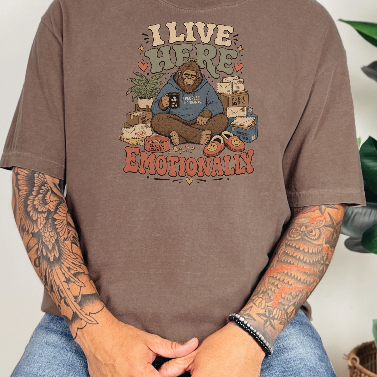 Bigfoot sitting with coffee mug, surrounded by mail and boxes, text "I LIVE HERE EMOTIONALLY".