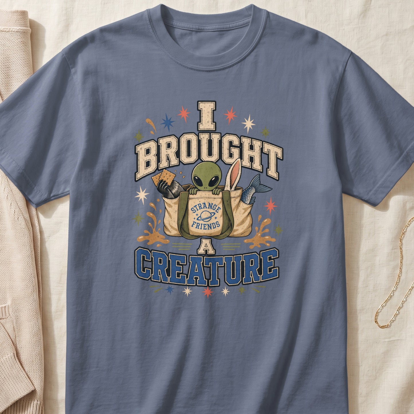 Blue t-shirt graphic with text "I BROUGHT A CREATURE" and an alien in a bag.