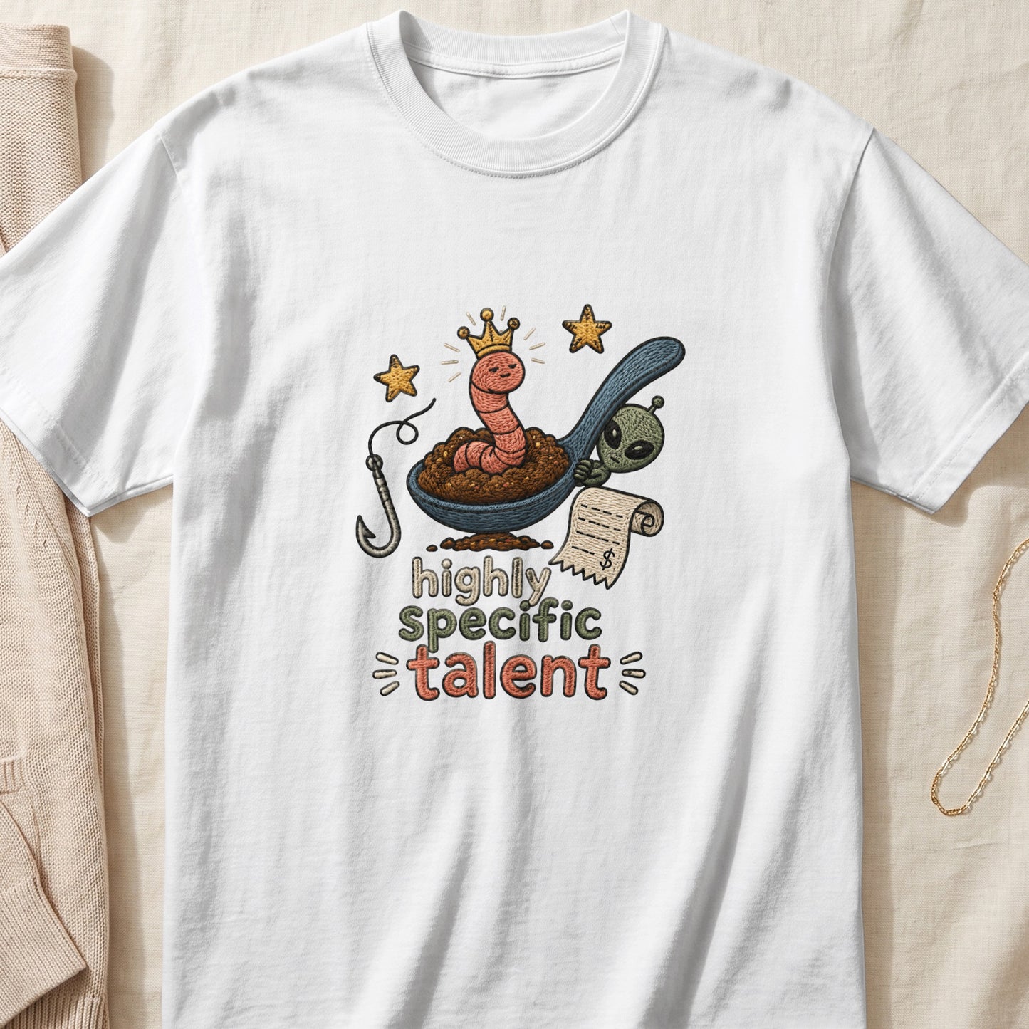 White t-shirt with embroidered graphic of worm in crown on spoon, alien, and text "highly specific talent".