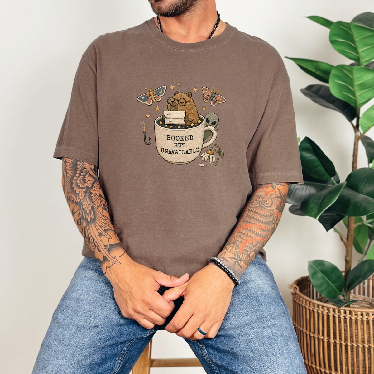 Brown t-shirt with "BOOKED BUT UNAVAILABLE" graphic featuring a capybara and alien.