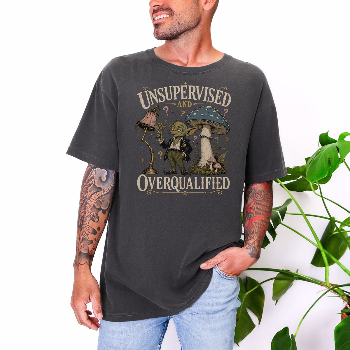Graphic tee with text "UNSUPERVISED AND OVERQUALIFIED" featuring a goblin and mushrooms.