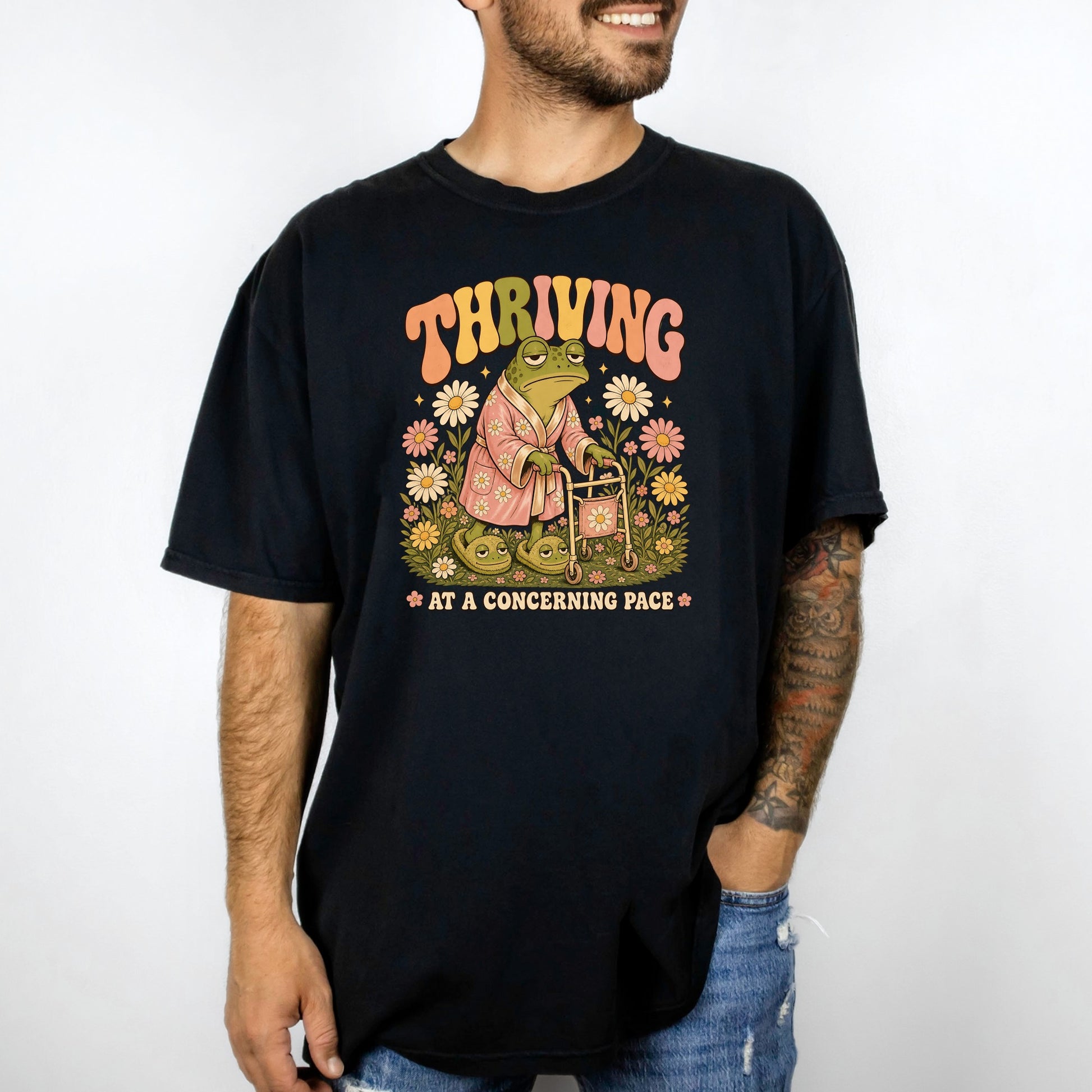 Black t-shirt with graphic of frog wearing robe, slippers, and walker, with text "THRIVING AT A CONCERNING PACE".
